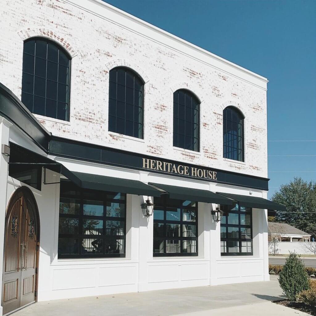 A white brick building with arched windows and black awnings, labeled 'Heritage House', stands under a clear blue sky in Tuscaloosa, Alabama.