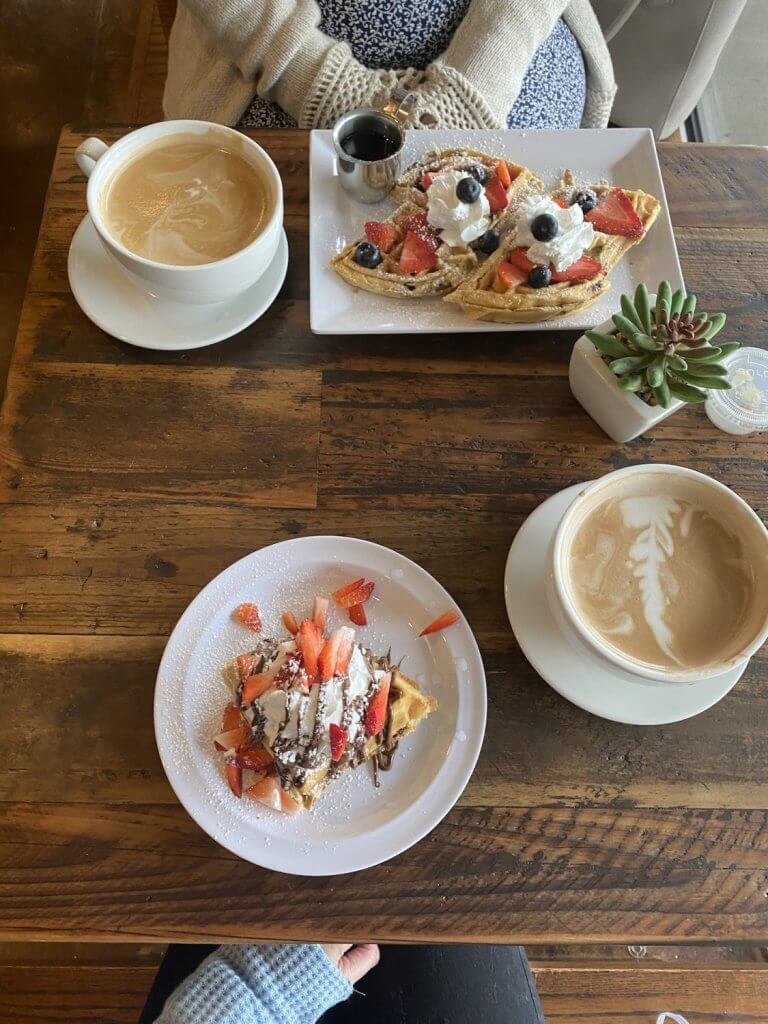 A cozy breakfast scene featuring a cup of latte art, a plate of waffles topped with fresh berries and whipped cream, and a smaller plate with a single waffle drizzled with chocolate and sprinkled with strawberries in Tuscaloosa, Alabama.