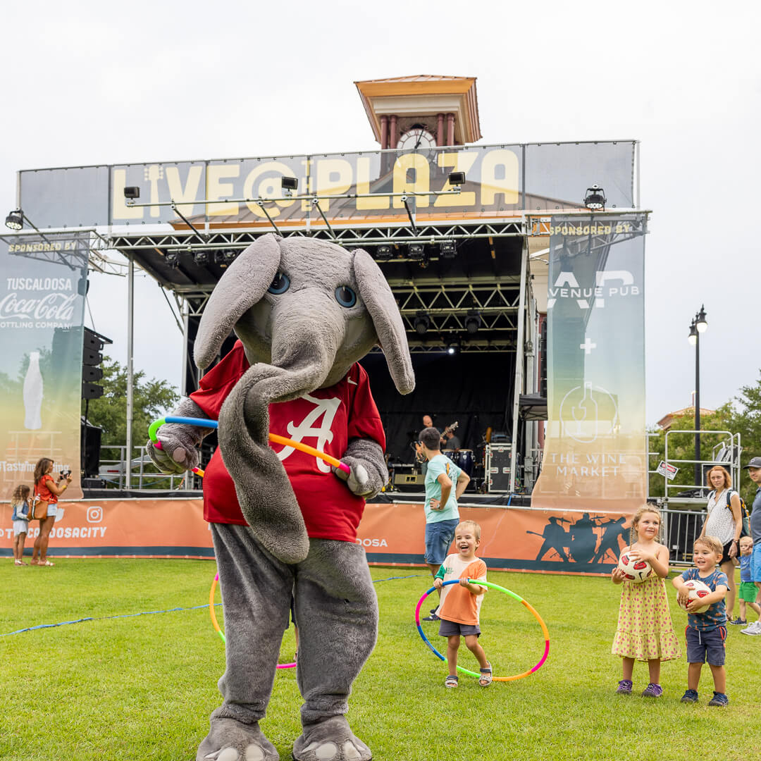 A lively outdoor event features a mascot elephant in a red 'A' shirt entertaining children with hula hoops on a grassy field in front of a stage in Tuscaloosa, Alabama.