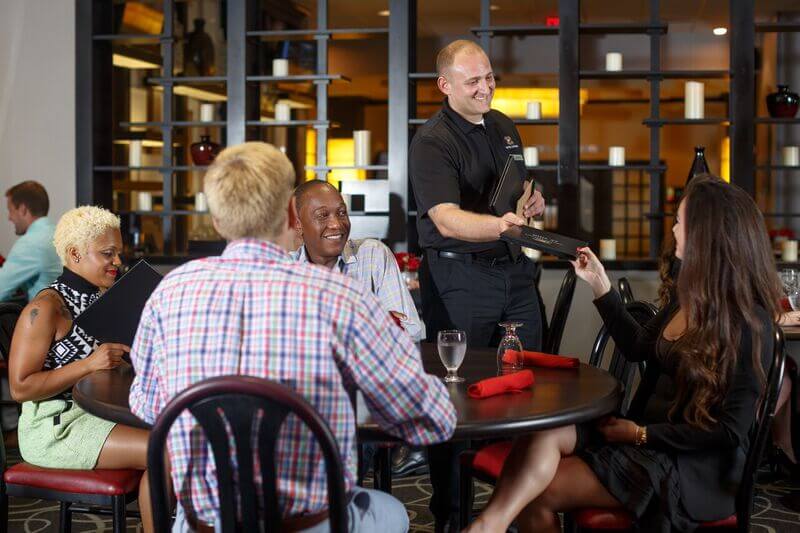 A group of people enjoying a meal together at a restaurant, with a waiter presenting a menu to a woman in Tuscaloosa, Alabama.