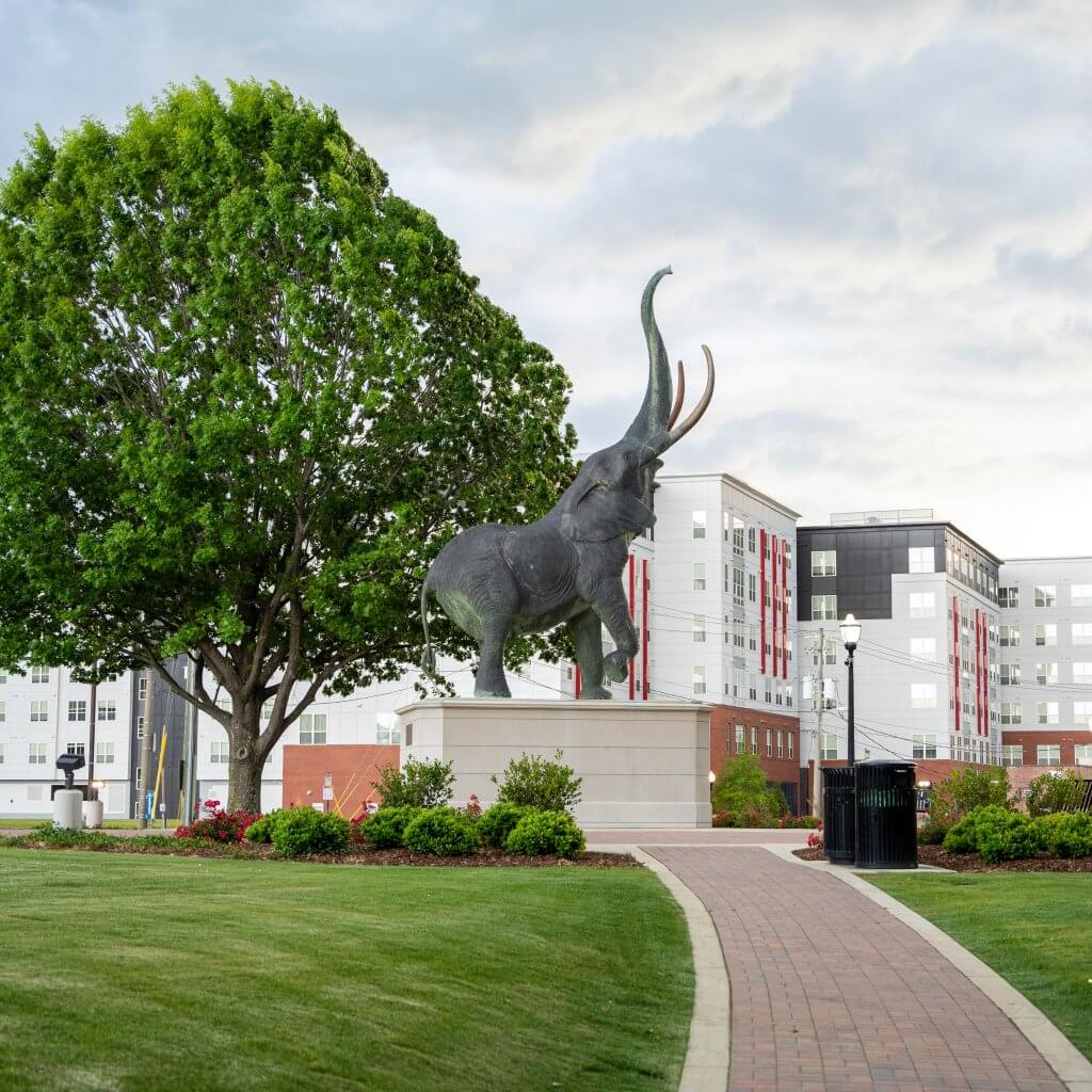 A majestic elephant statue stands proudly on a pedestal in a lush green park, with a towering tree and modern buildings in the background in Tuscaloosa, Alabama.
