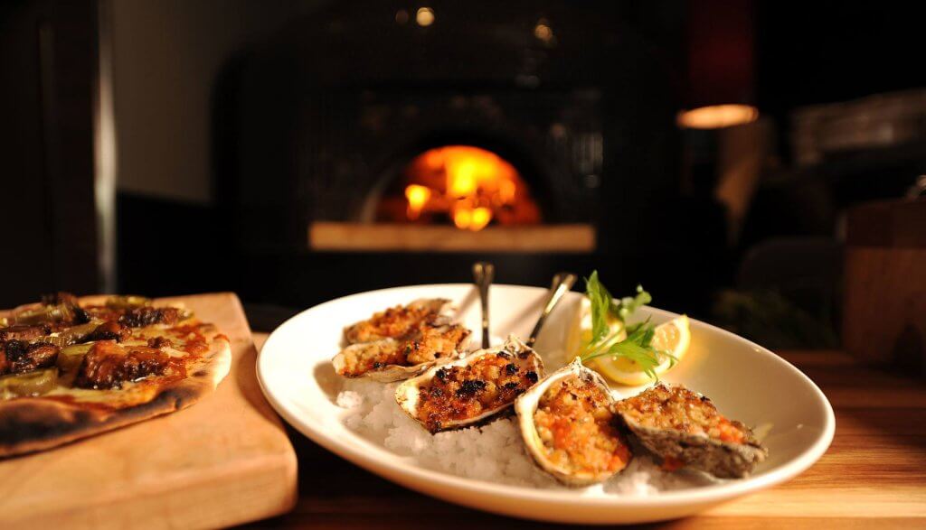 A plate of grilled oysters topped with a crispy breadcrumb mixture, served on a bed of ice with lemon wedges and fresh herbs, in front of a cozy fireplace in Tuscaloosa, Alabama.