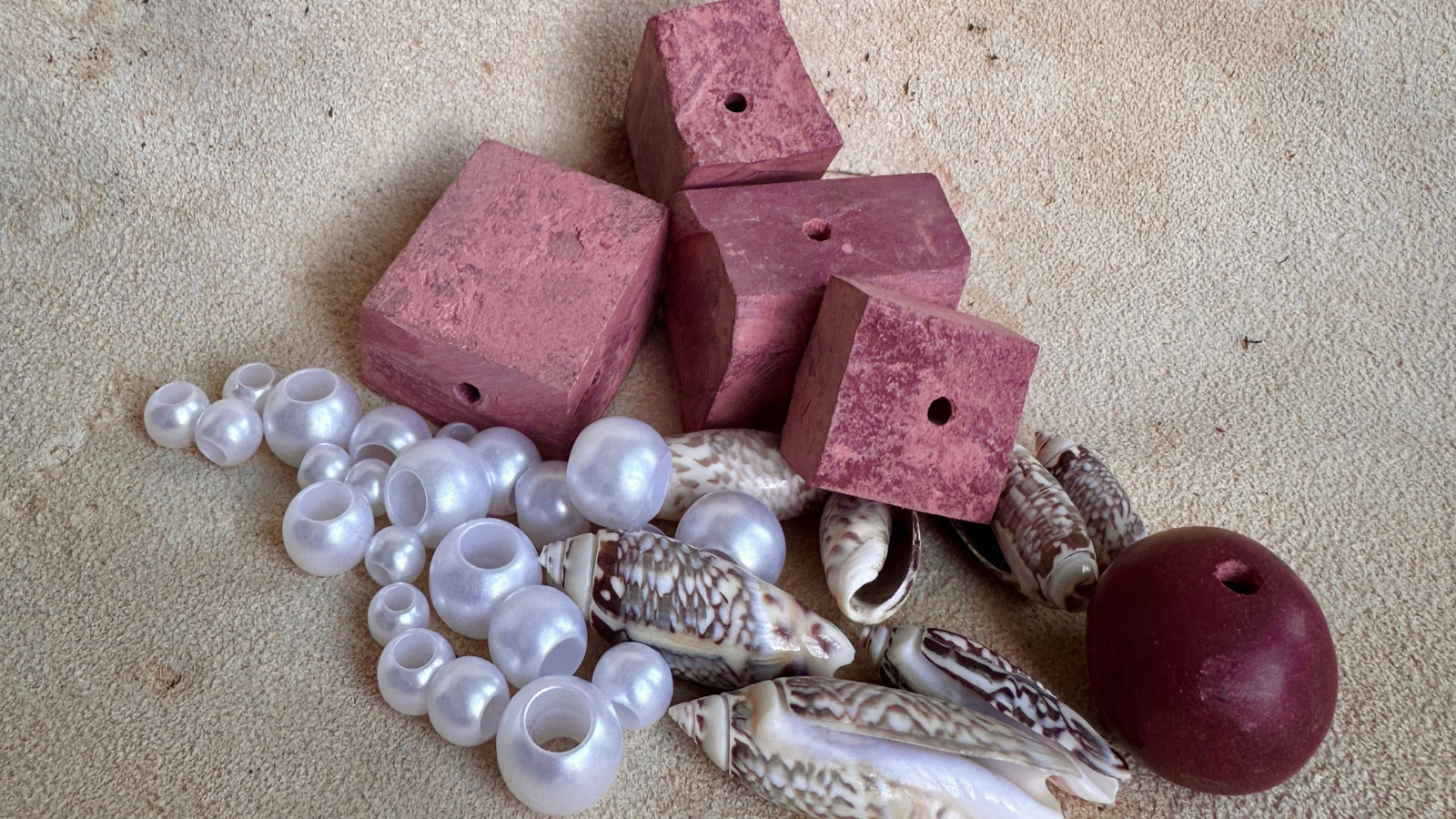 A collection of pink clay cubes, white and pink pearls, seashells, and a red stone on a sandy surface in Tuscaloosa, Alabama.