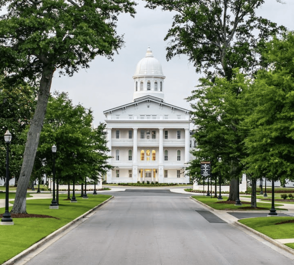A grand white building with a dome stands at the end of a tree-lined driveway in Tuscaloosa, Alabama.