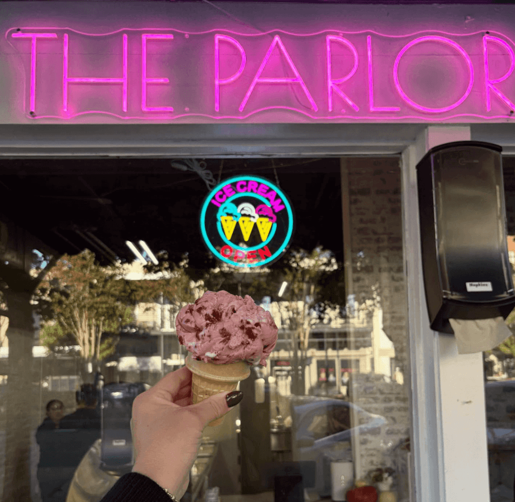 A hand with dark nail polish holds a pink ice cream cone outside The Parlor ice cream shop, with a neon sign and colorful ice cream logo visible through the window in Tuscaloosa, Alabama.