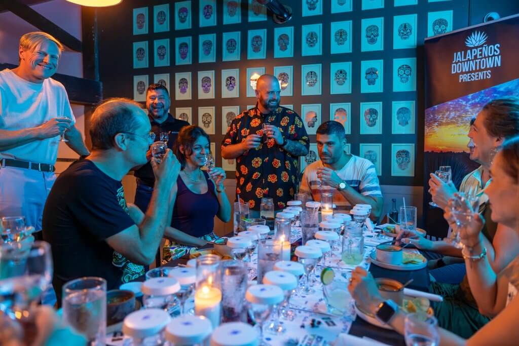 A lively group of people enjoys a tequila tasting event at Jalapenos Downtown, with a table full of shot glasses and a wall adorned with colorful sugar skull art in Tuscaloosa, Alabama.
