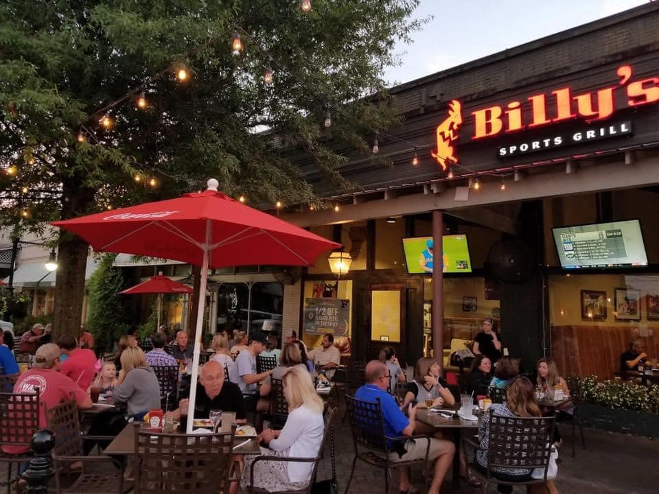 A lively outdoor patio at Billy's Sports Grill filled with people enjoying dinner and drinks under red umbrellas, with string lights and a large tree providing ambiance in Tuscaloosa, Alabama.