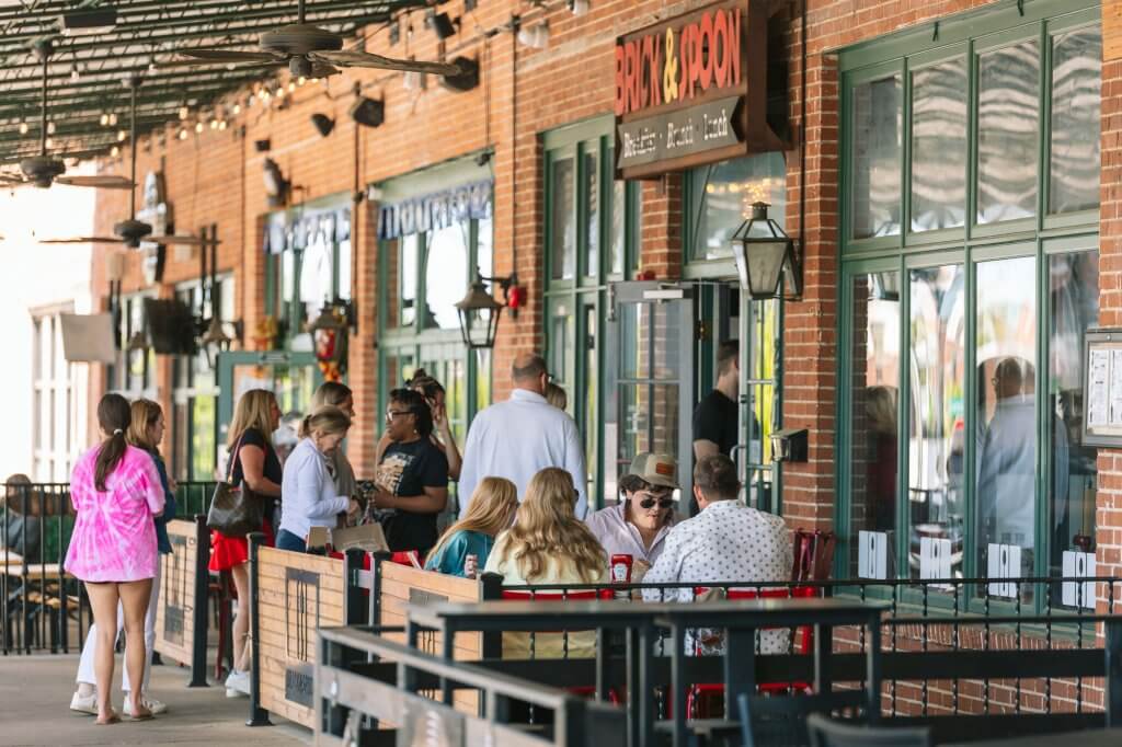A lively outdoor patio scene at a restaurant called Brick & Spoon, with patrons dining and socializing under a covered area adorned with string lights and ceiling fans in Tuscaloosa, Alabama.