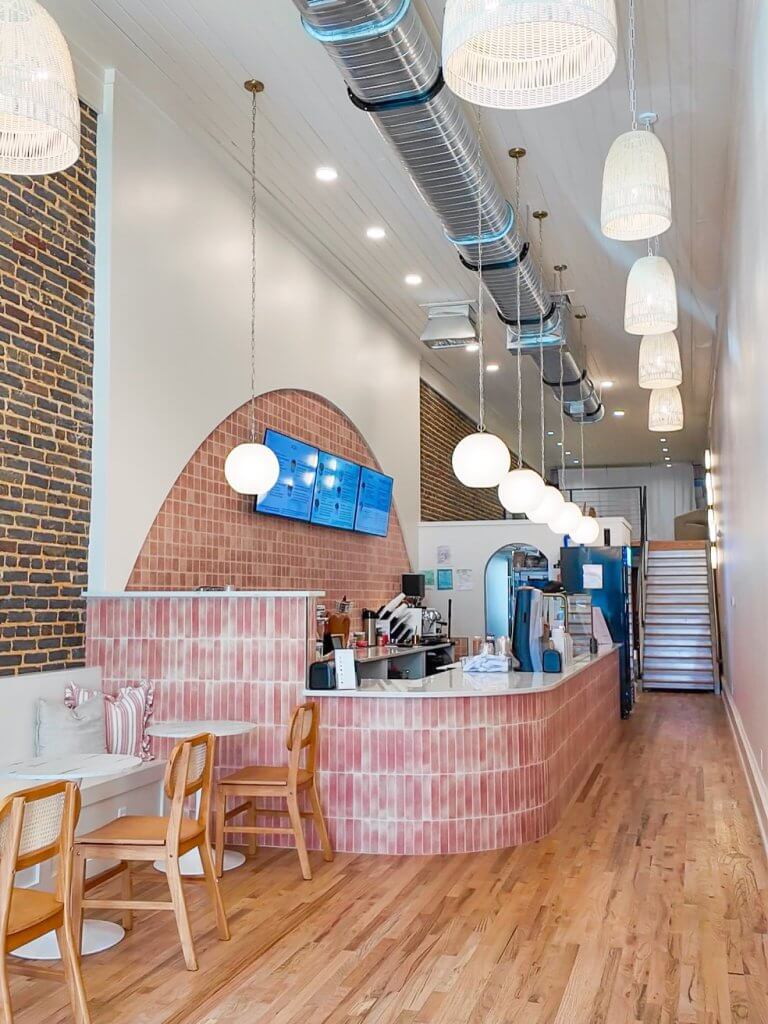 A modern cafe interior with a curved pink tiled counter, wooden tables and chairs, and hanging light fixtures in Tuscaloosa, Alabama.