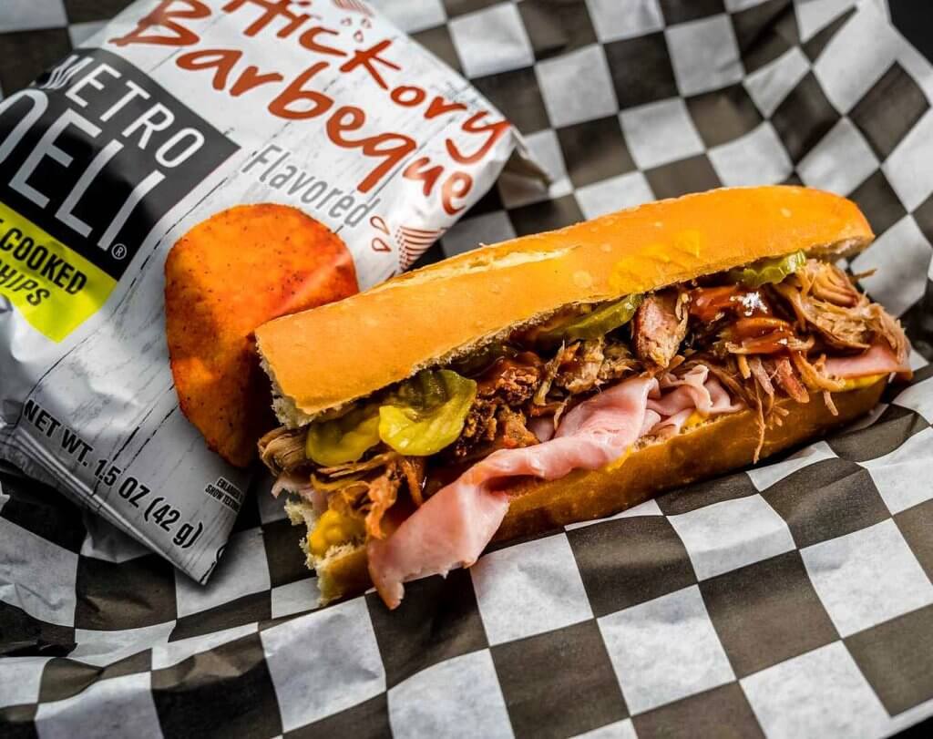 A hearty Cuban sandwich with pulled pork, ham, pickles, and mustard, served with a side of Hickory Barbecue flavored chips on a checkered paper wrapper in Tuscaloosa, Alabama.