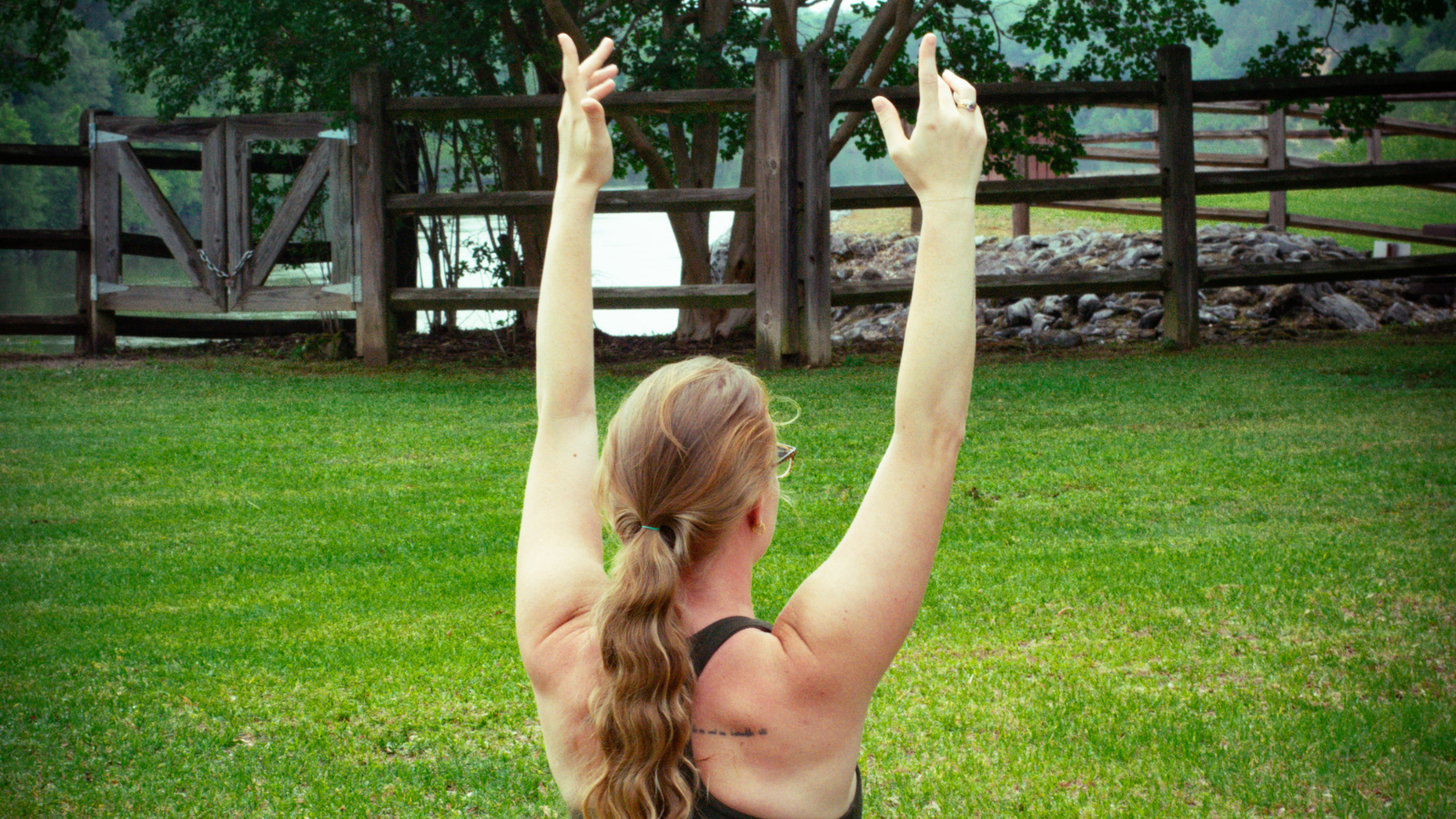 A woman with long hair in a ponytail raises her arms in a grassy field near a wooden fence in Tuscaloosa, Alabama.