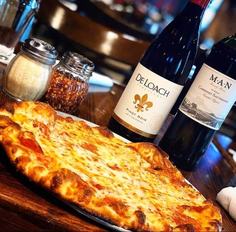 A freshly baked cheese pizza sits on a wooden table alongside two bottles of wine, a pepper shaker, and a Parmesan cheese shaker in Tuscaloosa, Alabama.