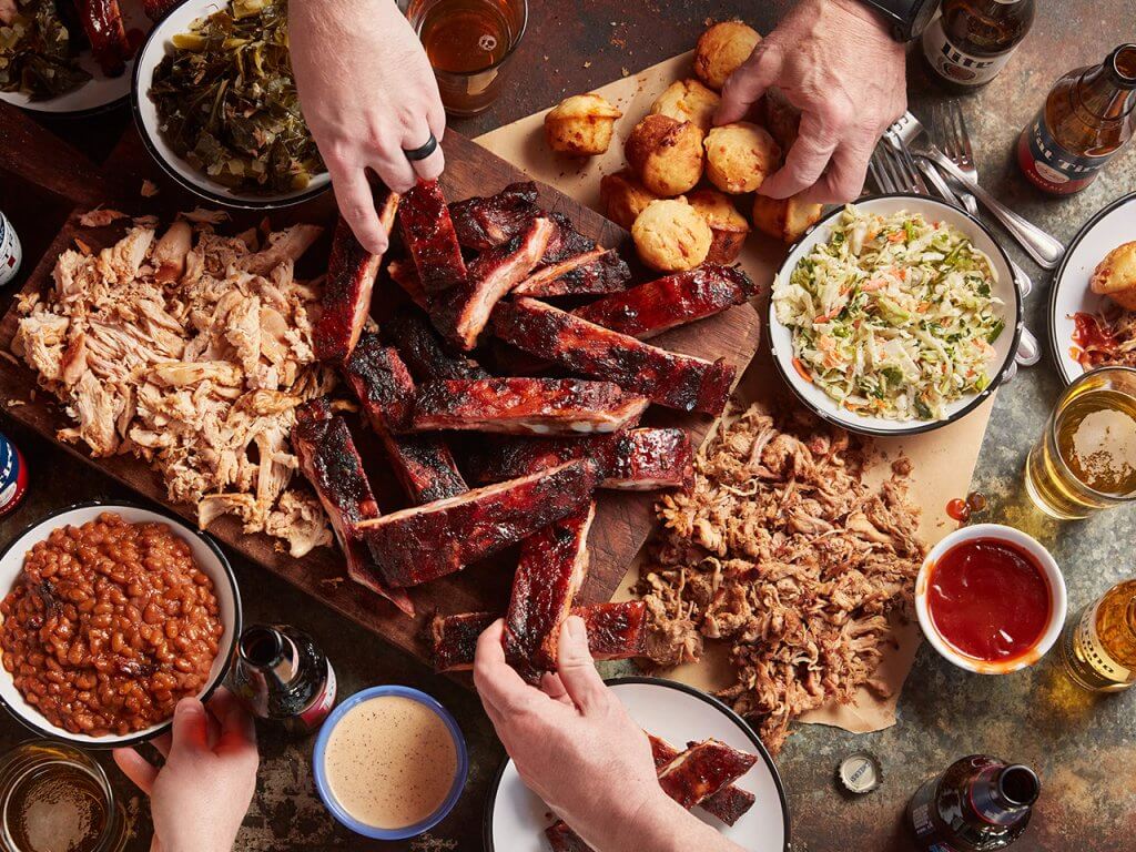A group of people gathered around a table laden with a variety of barbecue dishes, including ribs, pulled pork, baked beans, coleslaw, and cornbread, enjoying a meal together with drinks in hand in Tuscaloosa, Alabama.