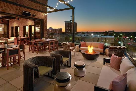 A rooftop terrace with a fire pit, comfortable seating, and dining tables overlooking a cityscape at sunset in Tuscaloosa, Alabama.