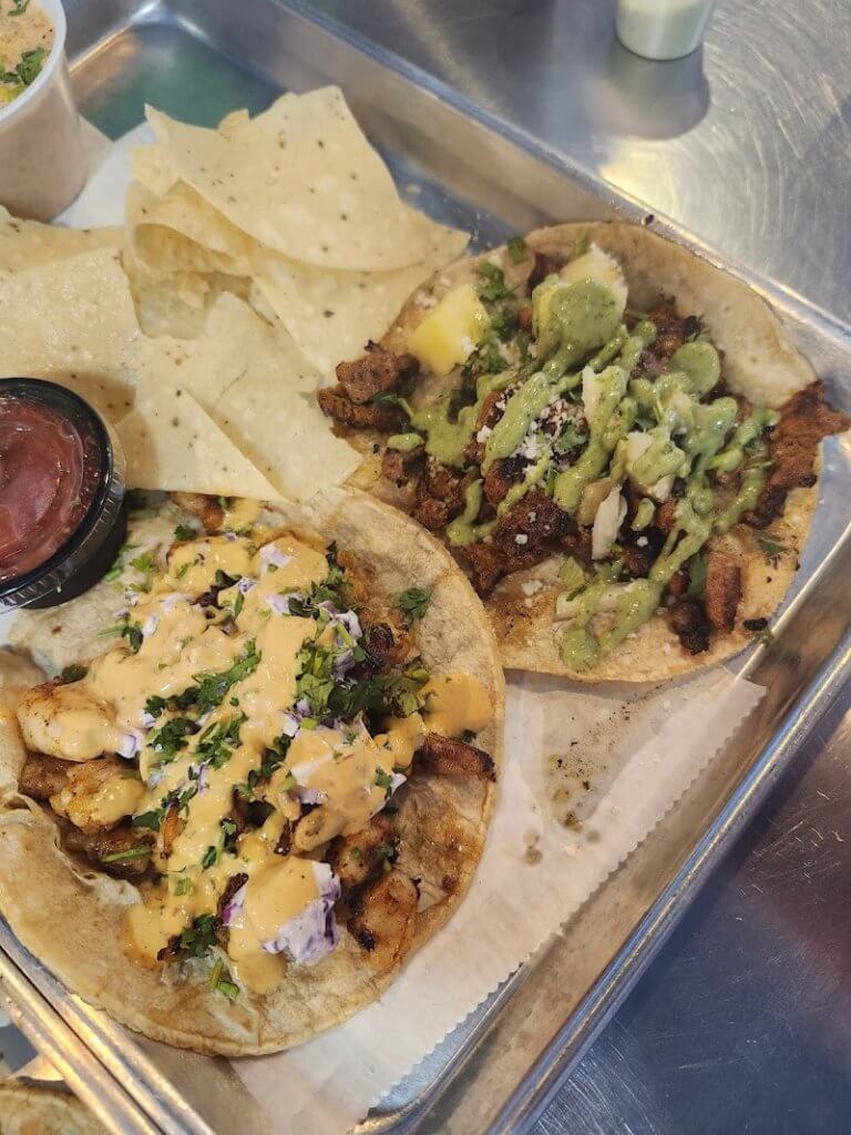 Two tacos with various toppings, including shrimp, meat, cheese, and sauces, served on a metal tray with tortilla chips and salsa in Tuscaloosa, Alabama.