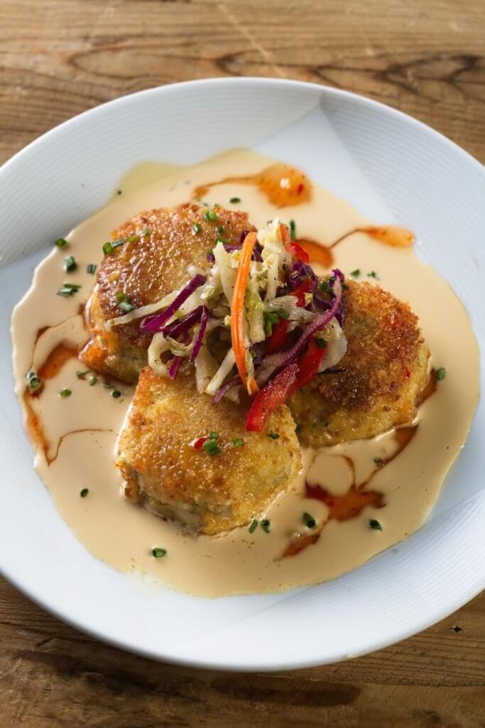 Three golden-brown crab cakes are artfully arranged on a white plate, topped with a colorful mix of shredded cabbage, carrots, and red peppers, and drizzled with a creamy sauce in Tuscaloosa, Alabama.