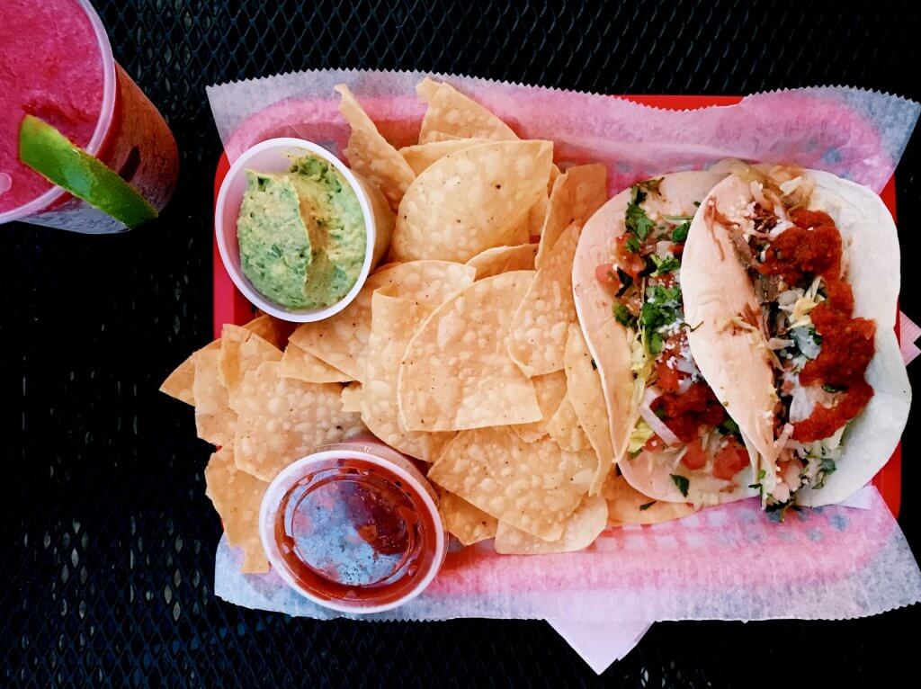 A vibrant meal featuring two tacos filled with meat, tomatoes, and cilantro, served with a side of tortilla chips, guacamole, and salsa, accompanied by a refreshing pink drink garnished with a lime wedge in Tuscaloosa, Alabama.