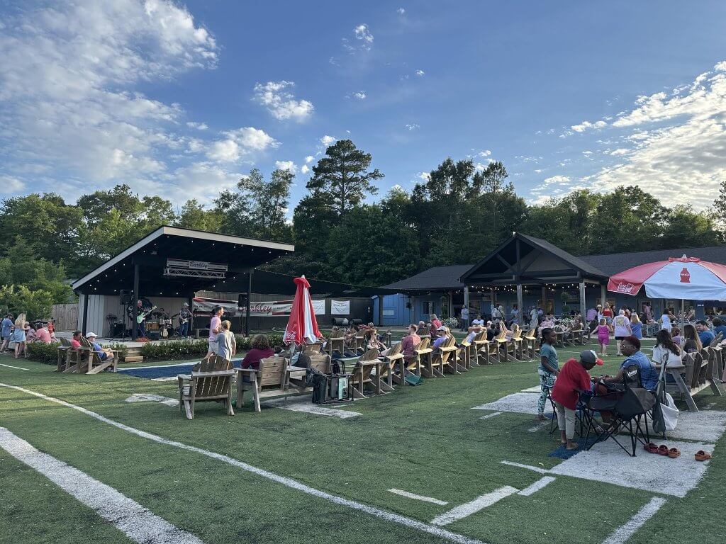 Outdoor concert on a grassy field with wooden bleachers, a stage, and a crowd enjoying the music under a clear blue sky in Tuscaloosa, Alabama.