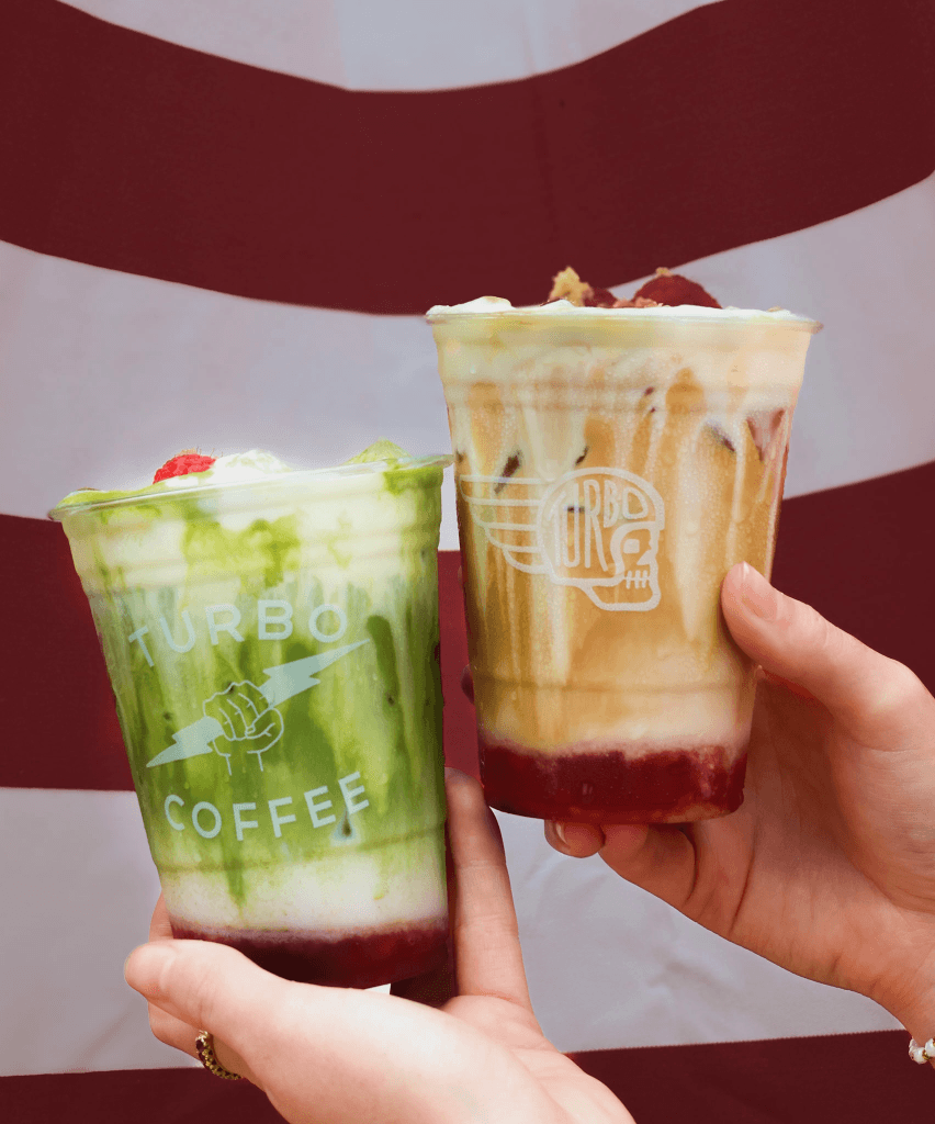 Two hands hold colorful Turbo Coffee drinks with unique designs against a red and white striped background in Tuscaloosa, Alabama.