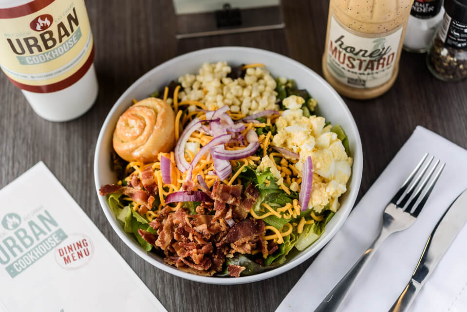 A colorful salad topped with bacon, cheese, corn, eggs, and onions, served with a honey mustard dressing and a dinner roll in Tuscaloosa, Alabama.