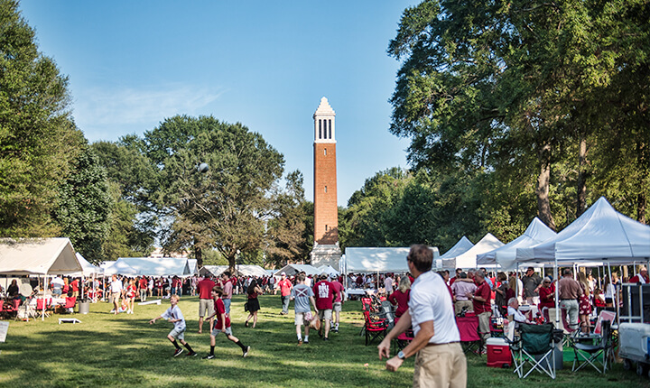 Alabama football fans tailgating on the Quad at the University of Alabama in Tuscaloosa, with tents, games, and Denny Chimes visible in the background.