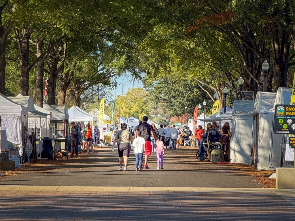 People walking through an outdoor art festival lined with white vendor tents under trees in Tuscaloosa, Alabama.