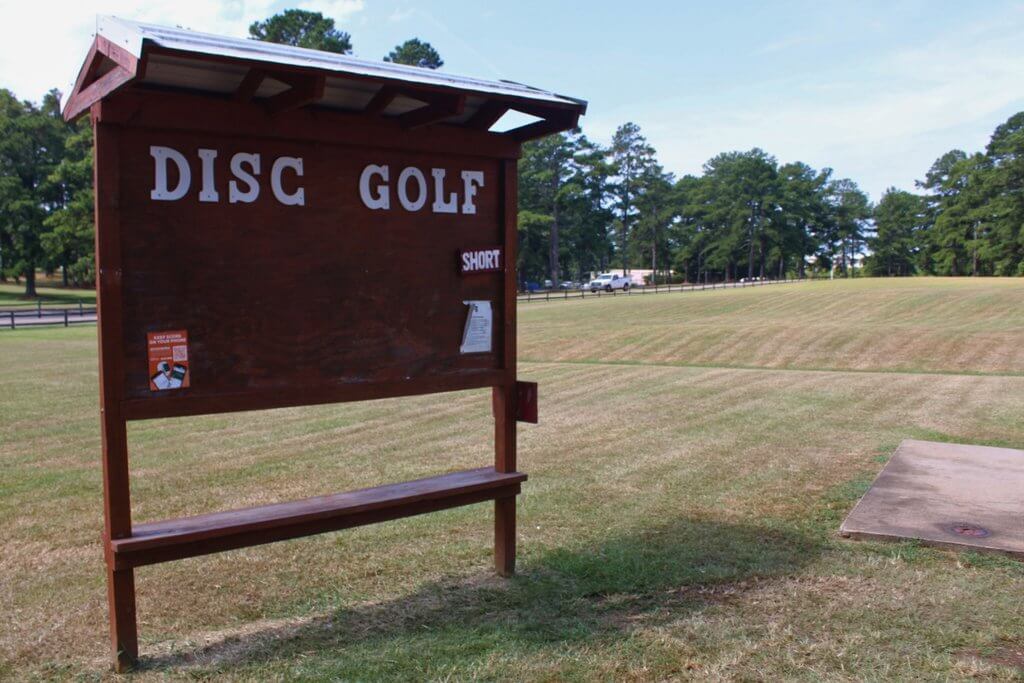 A wooden disc golf sign stands in a grassy field near a concrete tee pad in Tuscaloosa, Alabama.