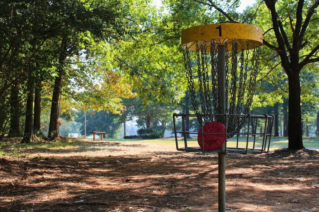 A red disc golf disc sits inside a metal basket on a sunny course in Tuscaloosa, Alabama.
