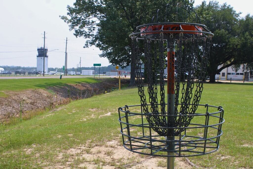 A disc golf basket stands on a grassy field near an airport control tower in Tuscaloosa, Alabama.