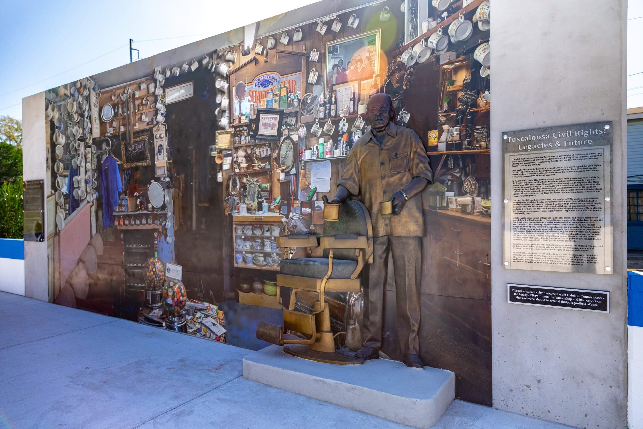 A bronze statue of a man stands before a mural of a barbershop in Tuscaloosa, Alabama.