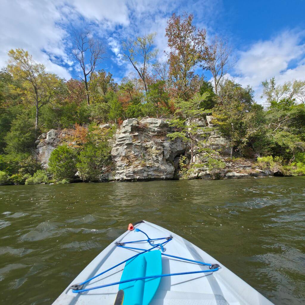 View from the bow of a kayak approaching a rocky cliffside covered in trees in Tuscaloosa, Alabama.