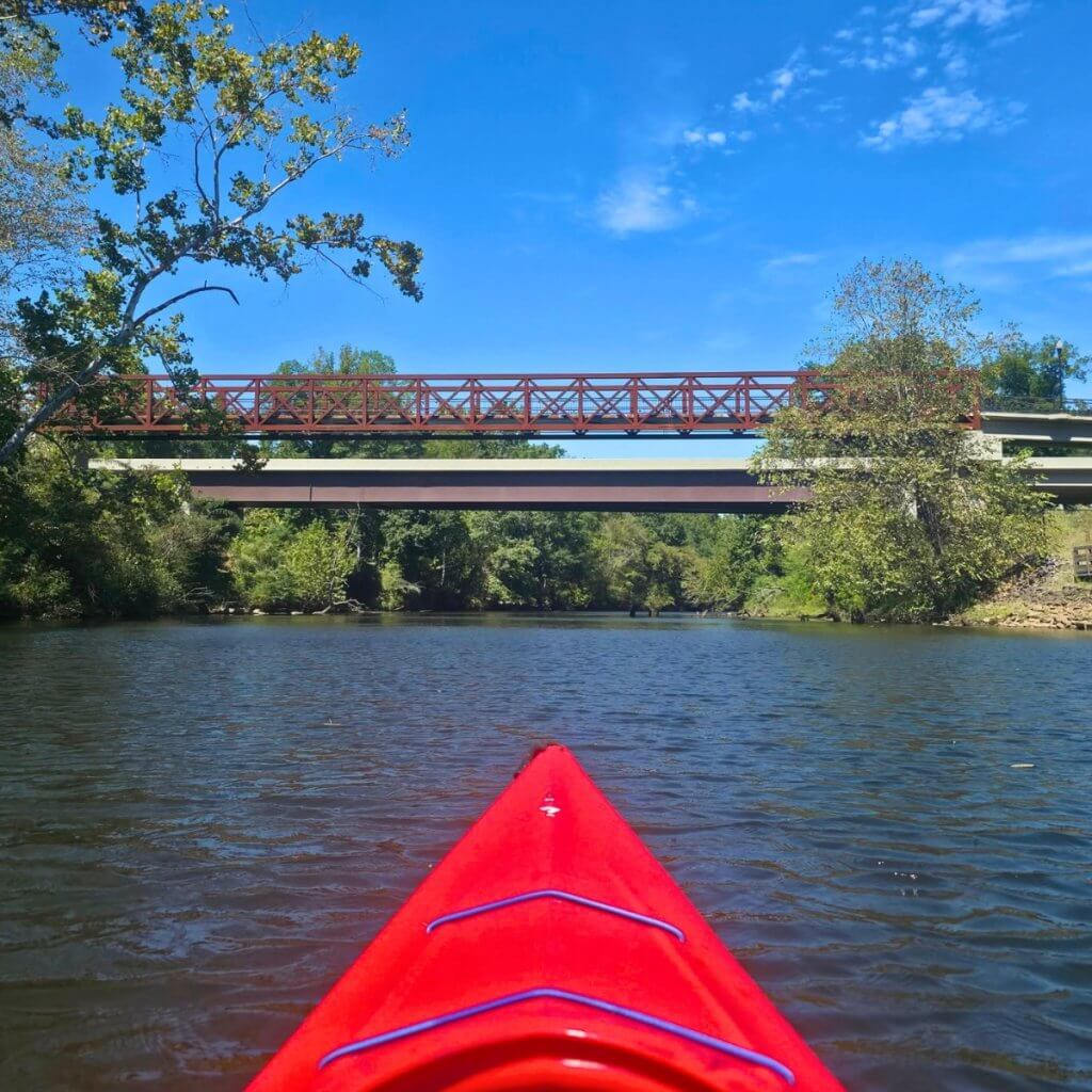 A red kayak floating on a calm river towards two bridges under a blue sky in Tuscaloosa, Alabama.