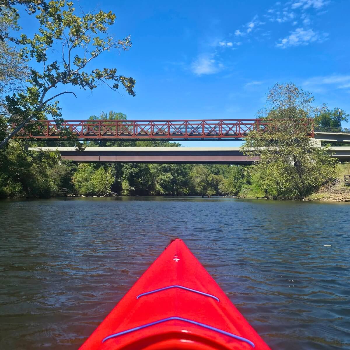 A red kayak floating on a calm river towards two bridges under a blue sky in Tuscaloosa, Alabama.